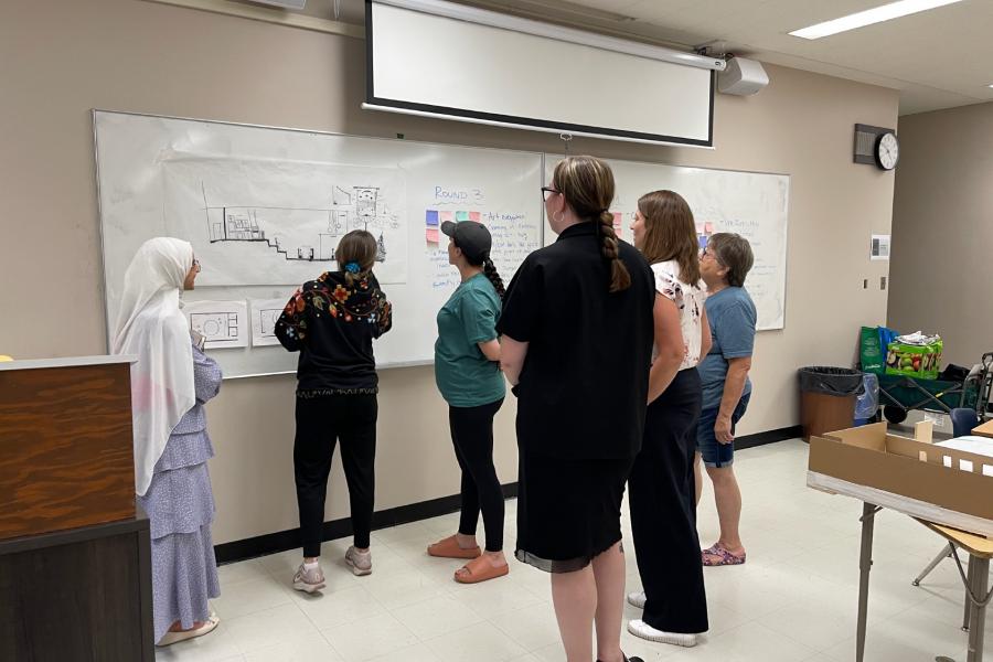 A group of six people stands in a classroom, attentively examining architectural drawings on a large whiteboard. The atmosphere is focused and collaborative.
