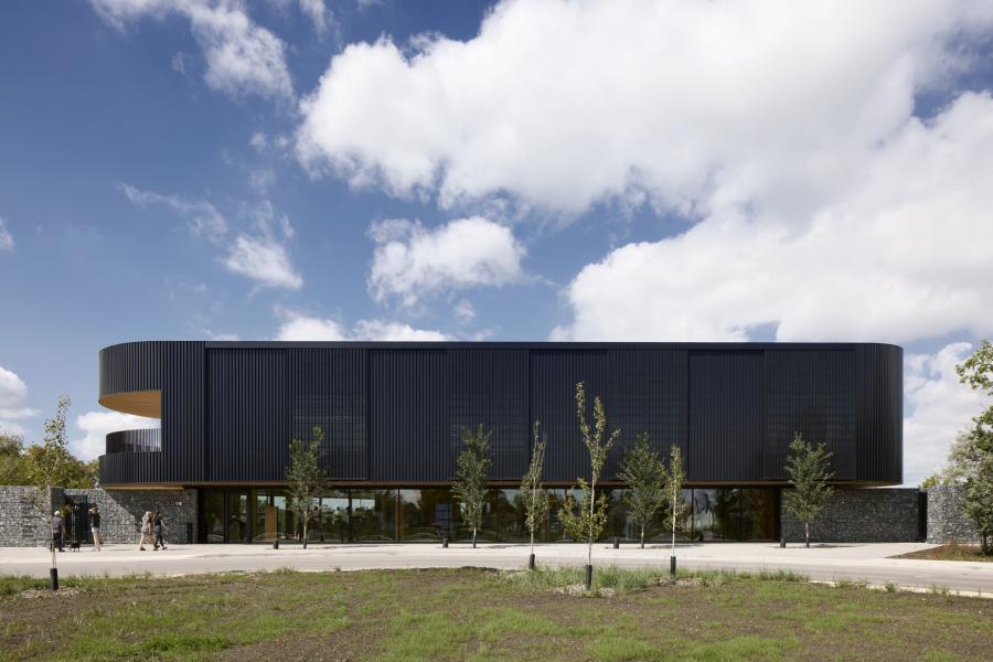 Modern building with a sleek, dark facade under a partly cloudy sky. Thin trees line the front, while a few people walk nearby. Calm and minimalist.