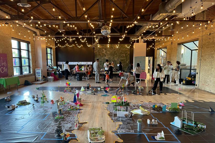 A spacious room with string lights and a wooden ceiling hosts a large art installation on the floor that resembles a cityscape. 