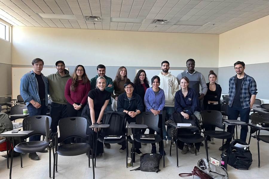 A group of fourteen diverse people in casual attire smile while standing and sitting in a classroom with desks and chairs.