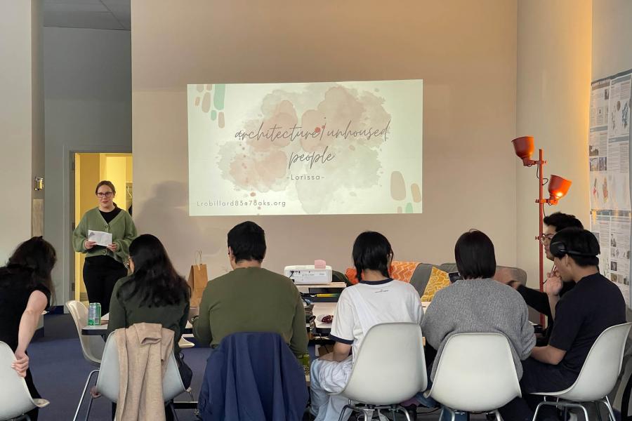 A woman is presenting to a group seated around a table, with a slide projected on the wall. 