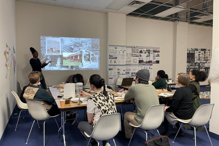 A woman presents architectural designs to a group seated around a table in a meeting room with blue carpet and white walls.