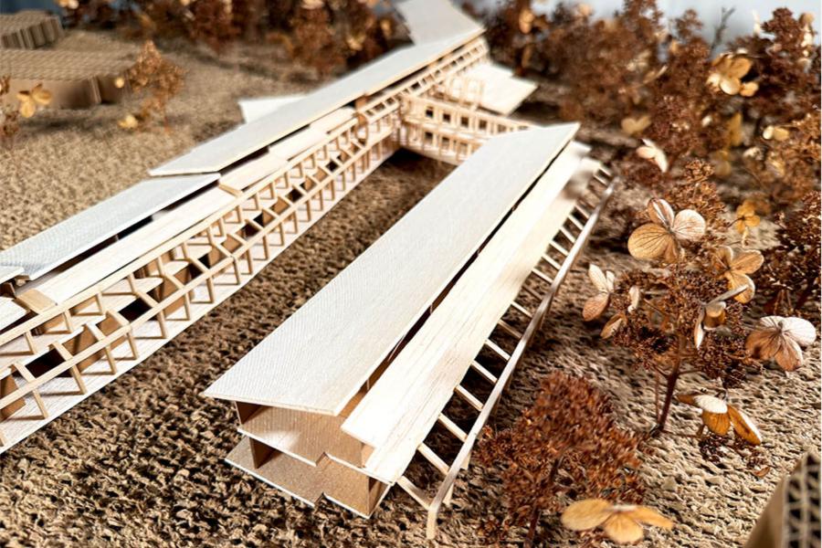 Architectural model with intersecting wooden structures on textured ground, surrounded by small brown trees, conveying a natural, rustic tone.
