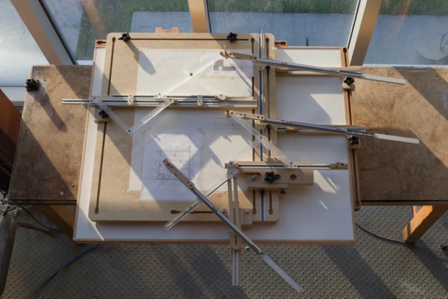A drafting table with a technical drawing under adjustment tools is bathed in soft, warm sunlight.