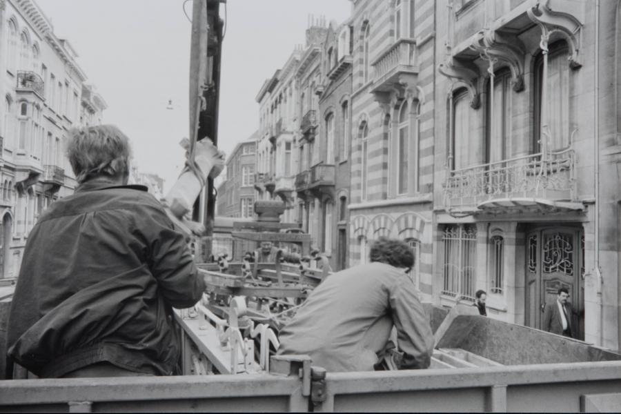 Black-and-white street scene: two men ride in an open truck past ornate historic buildings, evoking a busy, gritty urban atmosphere.