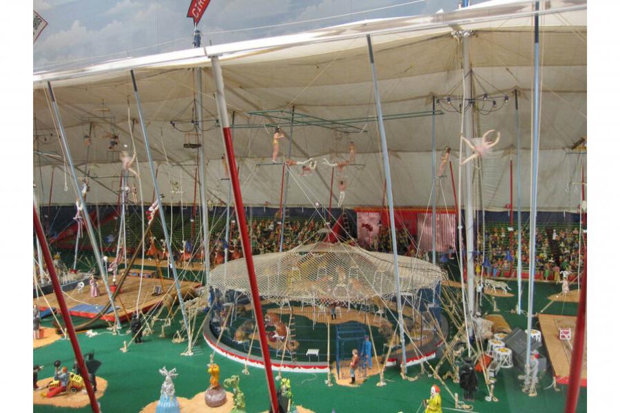Colorful miniature circus under a white tent, with suspended trapeze artists, a safety net, crowds, and carnival figures creating a lively scene.