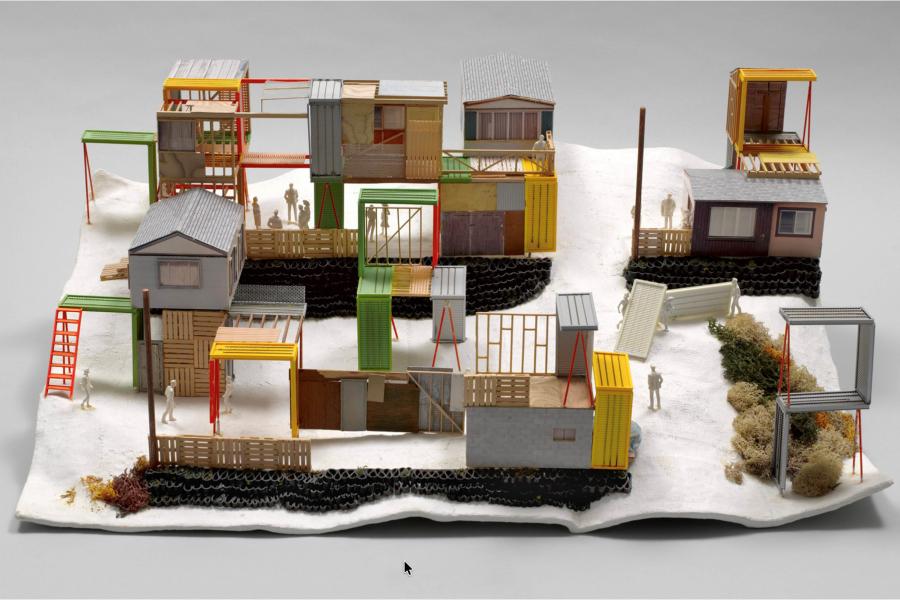 Miniature architectural model of a colorful stacked settlement on a white base, with porches, ladders, railings, shrubs, and tiny figures.