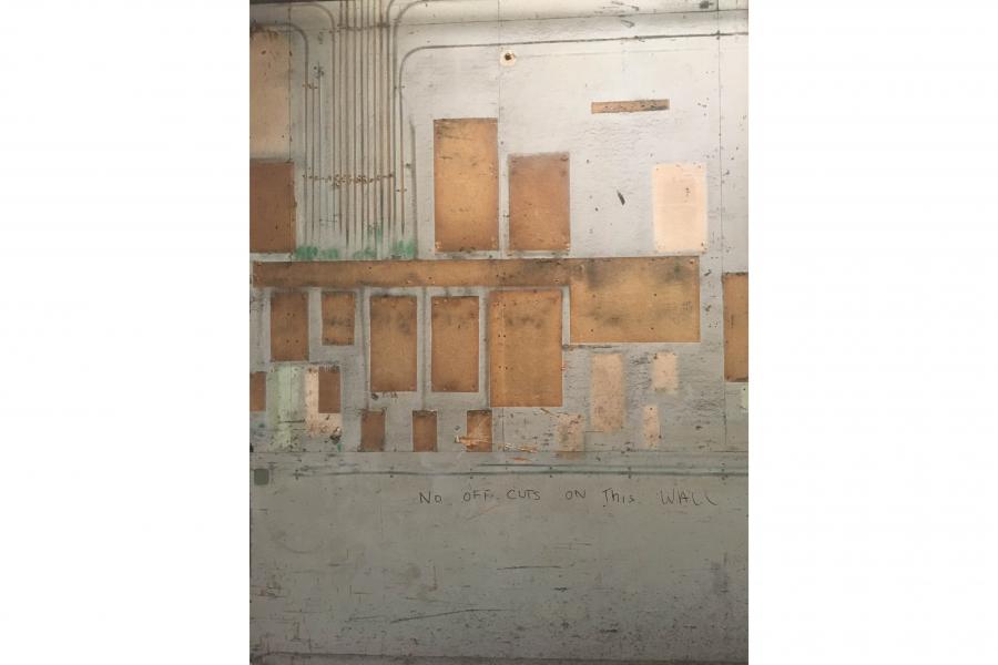 Weathered industrial wall with exposed electrical conduits, boarded patches, faded paint, and faint graffiti, creating a stark, neglected mood.