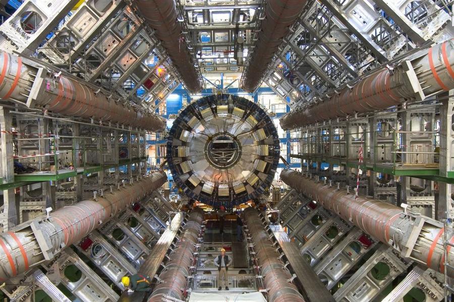 Interior of a massive particle collider detector, with concentric machinery, cables, pipes, and steel structures creating a futuristic, technical scene.