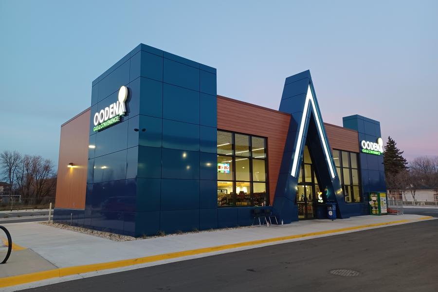 A modern OODENA convenience store with blue glass towers, a red-brown facade, and a striking triangular entrance stands beside an empty road at dusk.