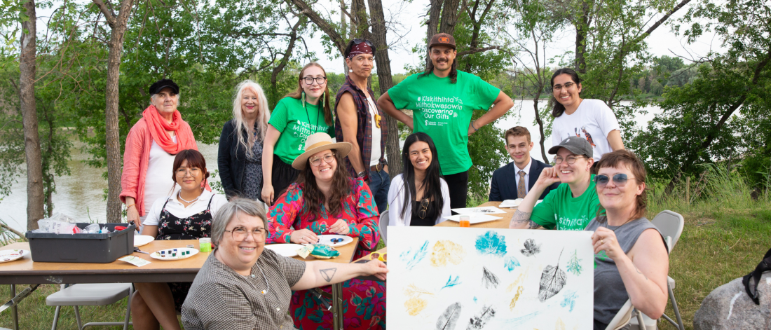 a group of people stand and sit around a table in a grassy field, smiling towards the camera displaying prints they created in the workshop