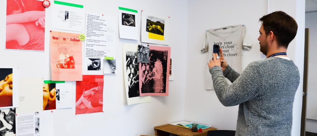 A person photographing a wall of expressive black-and-white and pink-toned prints, drawings, and text-based art.