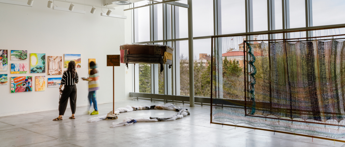 Two viewers explore a student exhibition featuring paintings, sculpture, and installation in a sunlit ARTlab studio.