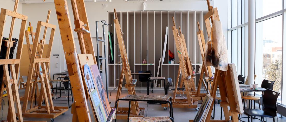 A bright painting studio at the School of Art with large windows, wooden easels, paint-splattered stools, and works in progress arranged throughout the space.