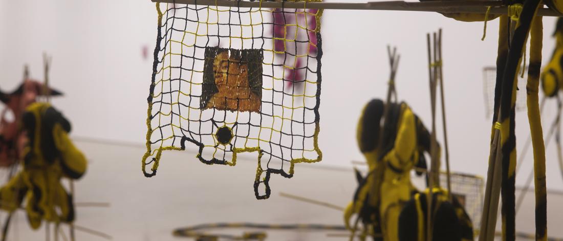 A suspended woven textile square of yellow-and-black cord with an image of a person’s face in the middle, hanging on a wooden support in front of other soft sculptures in the gallery.