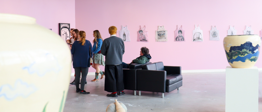 Visitors gathered in a gallery with pink walls, viewing drawings and a large ceramic vessel, with seating in the center.