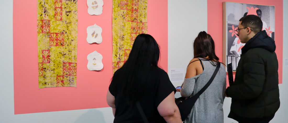 Visitors view mixed-media artworks on a pink wall, reading the accompanying text.