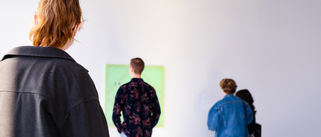 Wide shot of three people viewing artwork in a bright, white gallery; a person with a ponytail in the foreground faces a soft green painting, while two others stand to the right observing nearby works.