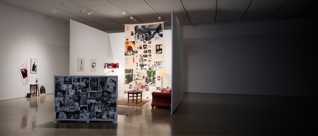 Gallery installation featuring a domestic living-room setting with family photographs, textiles, furniture, and a floor-to-ceiling collage exploring memory and personal archives.