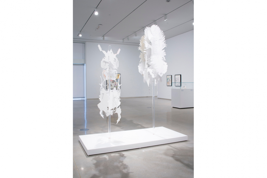 Two abstract 3d form, made of intersecting cut out white paper hand from the ceiling in the middle of a room, over a white low platform.