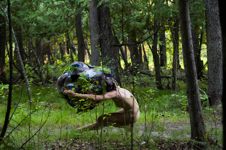 Located in a treed forest, a female figure in a nude leotard dances while holding a large boulder object, made of fabric