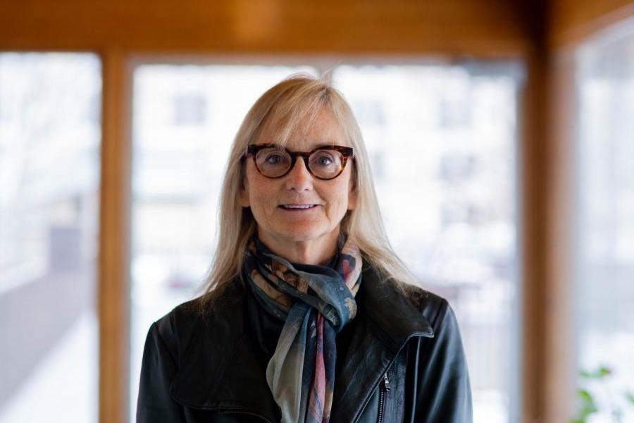 Liv Valmestad face the camera directly, smiling with long blonde hair, dark rimmed glasses, and a green scarf.