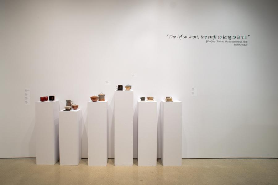 A display of ceramic works on white pedestals with a quote on the wall above: "The lyf so short, the craft so long to lerne."