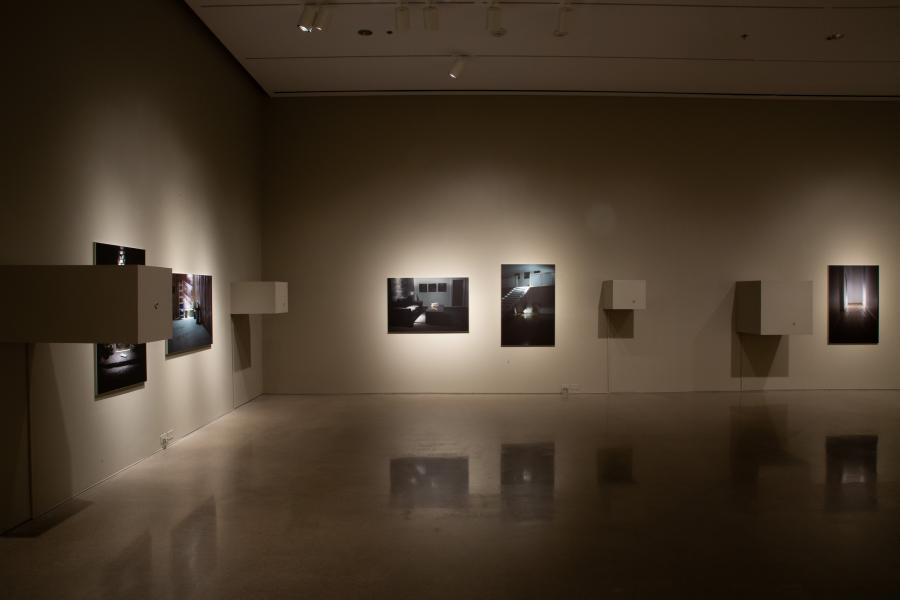 A close-up of a gallery wall featuring black-and-white photographic prints and minimalist wall-mounted sculptural boxes with small openings.
