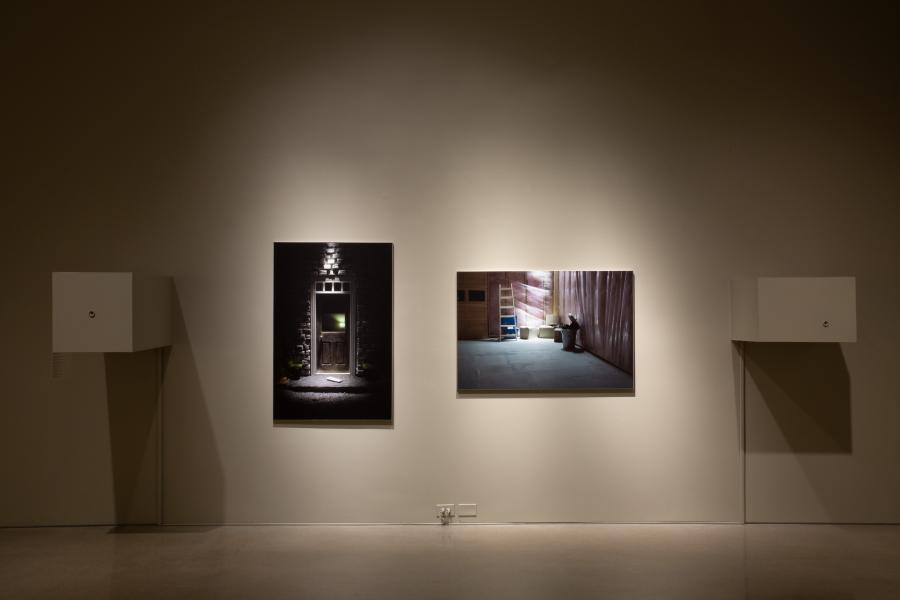 One image features an illuminated exterior door with a book on the step, and the other depicts an interior scene with construction materials and a pink plastic sheeting backdrop. Two small white boxes are mounted on the wall beside each photograph.