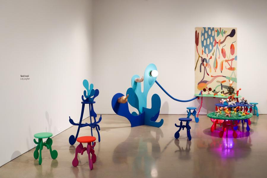 A wide view of the installation with colorful stools, abstract sculptural forms, and a vibrant wall tapestry in the background.