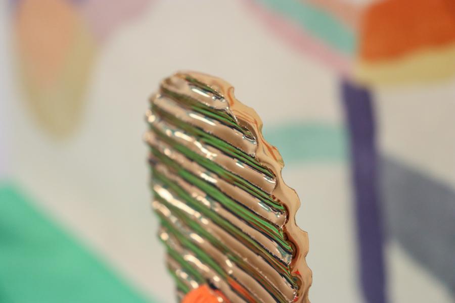 A detailed close-up of a metallic, ridged ceramic piece with an orange base, set against a blurred abstract background.