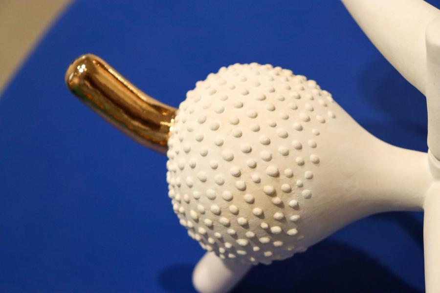 A white, textured ceramic piece with gold accents, resembling a hybrid between organic and abstract forms, displayed on a blue surface.