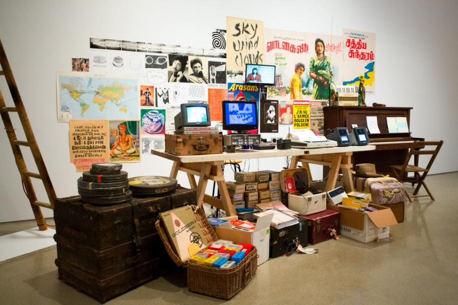A close-up of a curated table filled with objects like film reels, old typewriters, vintage posters, books, and boxed archives, creating a dense, layered installation.