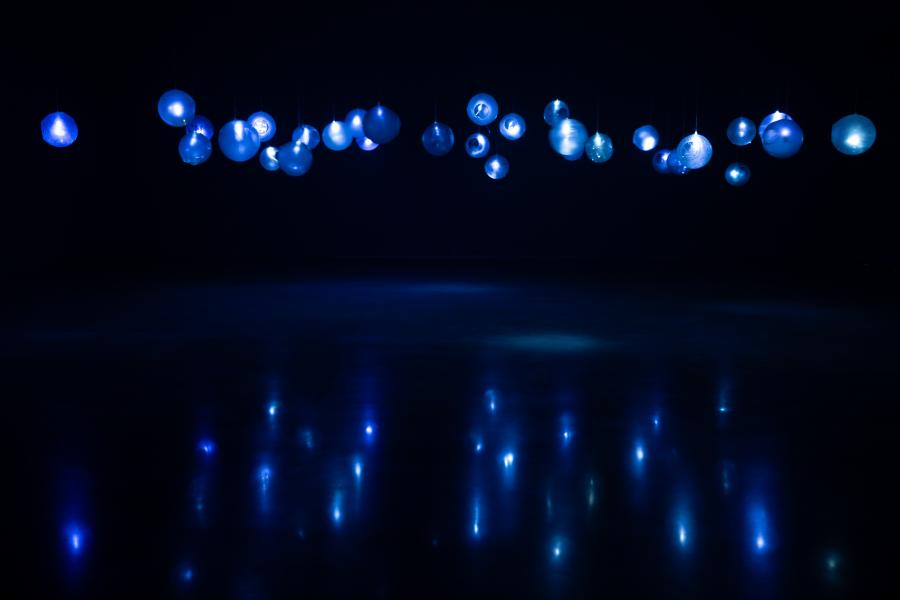 A dark gallery space with numerous glowing blue glass orbs suspended from the ceiling, reflecting their light on the polished floor below.