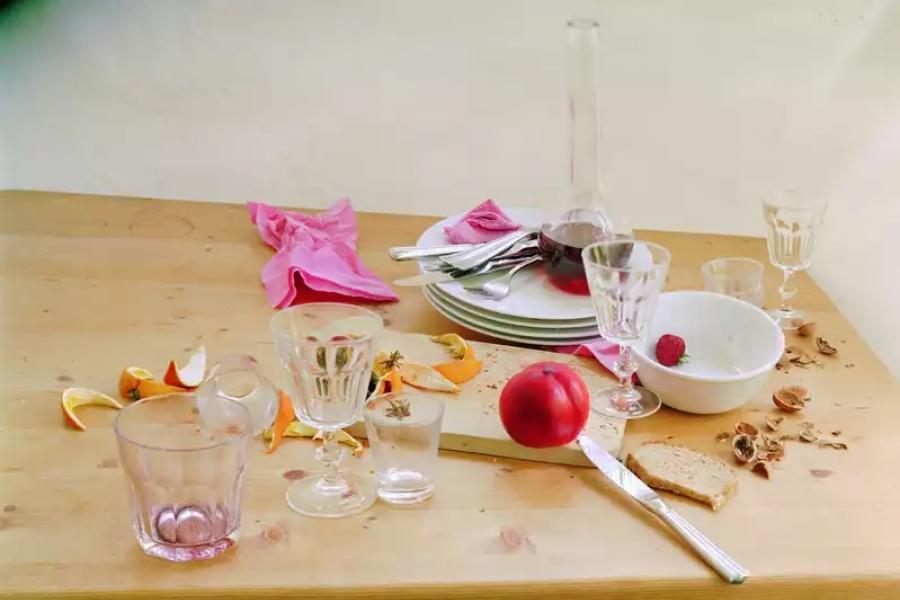 A still life photograph of a wooden table set with used dishware and remnants of a meal. The scene includes empty glasses, a carafe with red liquid residue, a plate stack with utensils and pink napkins, a sliced peach, orange peels, a single raspberry in a white bowl, and scattered crumbs. The overall scene suggests an intimate moment after a meal.