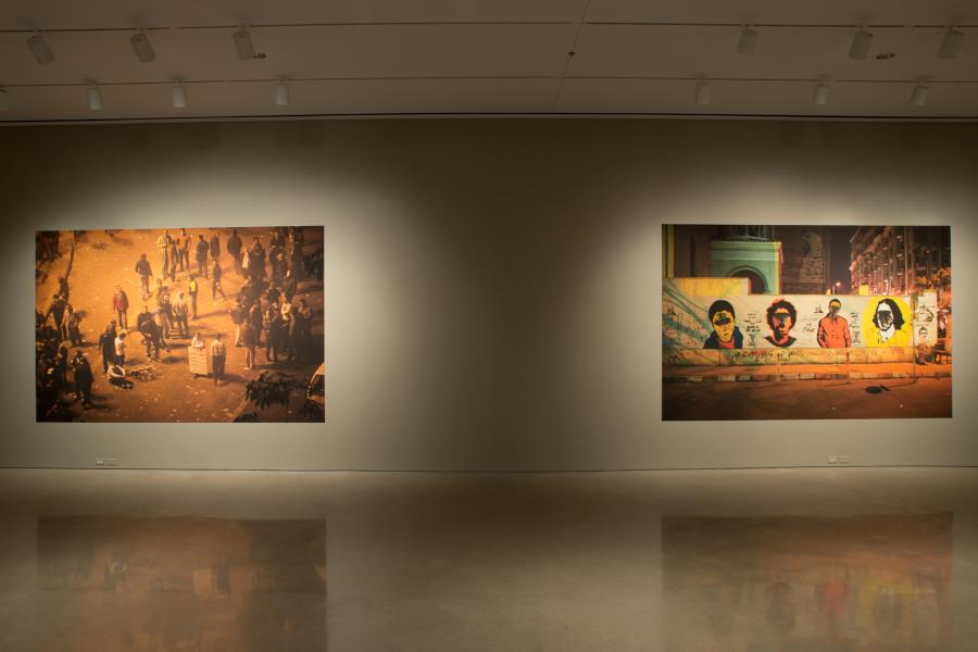 Two large-scale photographs displayed side by side in an art gallery. The left image depicts a crowd gathered in a nighttime street scene, illuminated by warm lighting. The right image showcases a wall with graffiti portraits and vibrant yellow and red hues, likely taken in an urban environment.