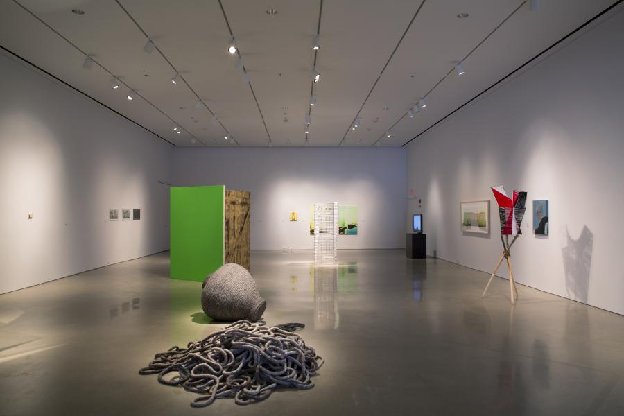 A contemporary gallery installation featuring diverse artworks, including a green panel, sculptures, and a rope sculpture on the polished concrete floor.