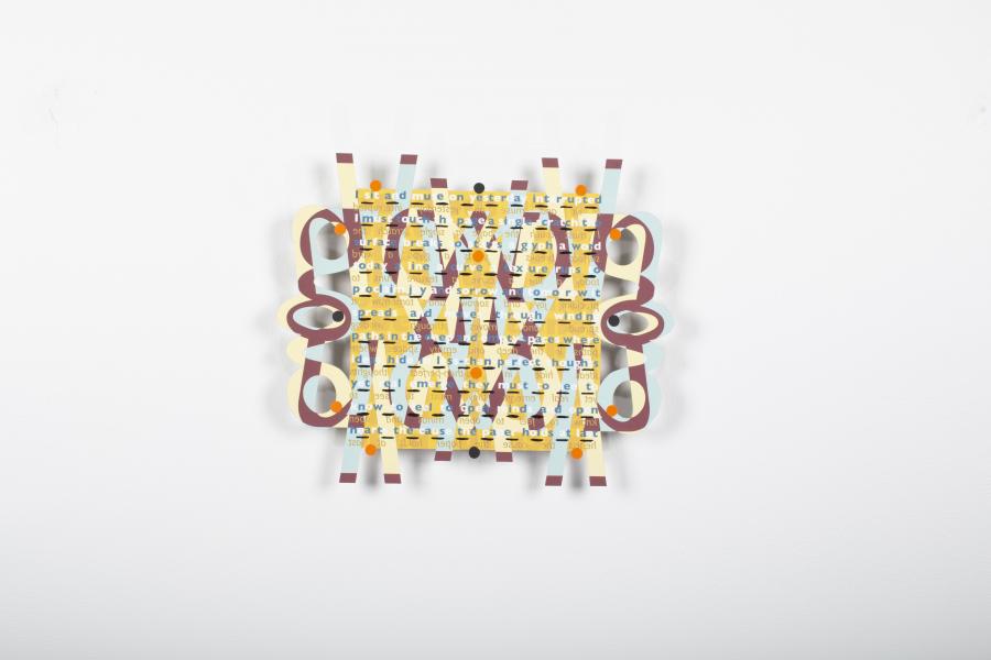 A colourful abstract artwork mounted on a white wall. The piece features intricate interwoven patterns with text, geometric shapes, and cutouts. The design includes shades of yellow, maroon, blue, and orange, creating a layered, dynamic composition that combines textual and visual elements. The symmetrical layout and bold colours make it visually engaging and balanced.