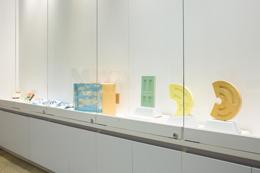 A well-lit glass display case showcasing objects. Items include ceramic sculptures in pastel hues, a blue translucent sculpture with cloud motifs, and a bright green door-shaped piece. Each item is displayed on a minimalist white base.