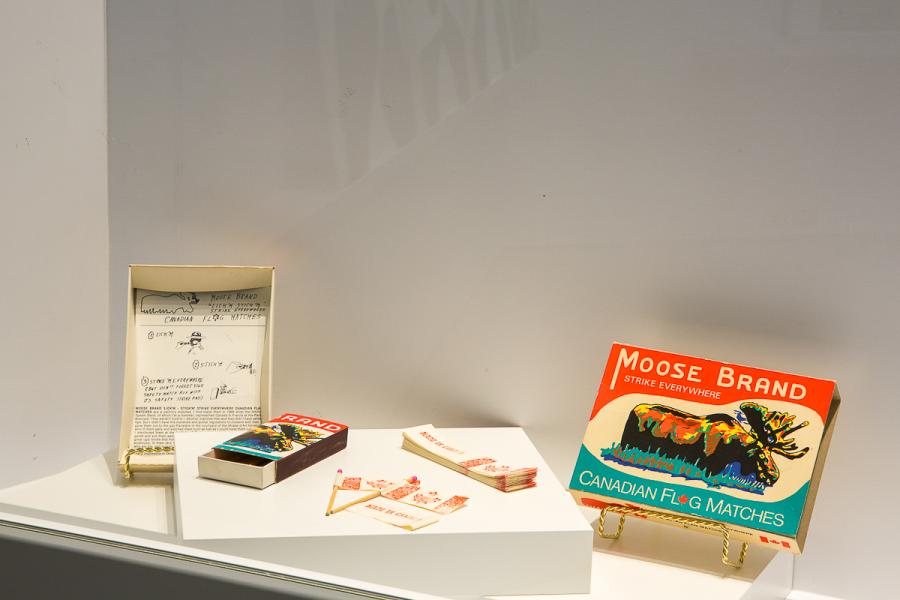 A close-up of a display case showing vintage items. Included are a matchbox with the label "Moose Brand - Canadian Flag Matches," accompanying red maple leaf matchsticks, and related historical sketches.