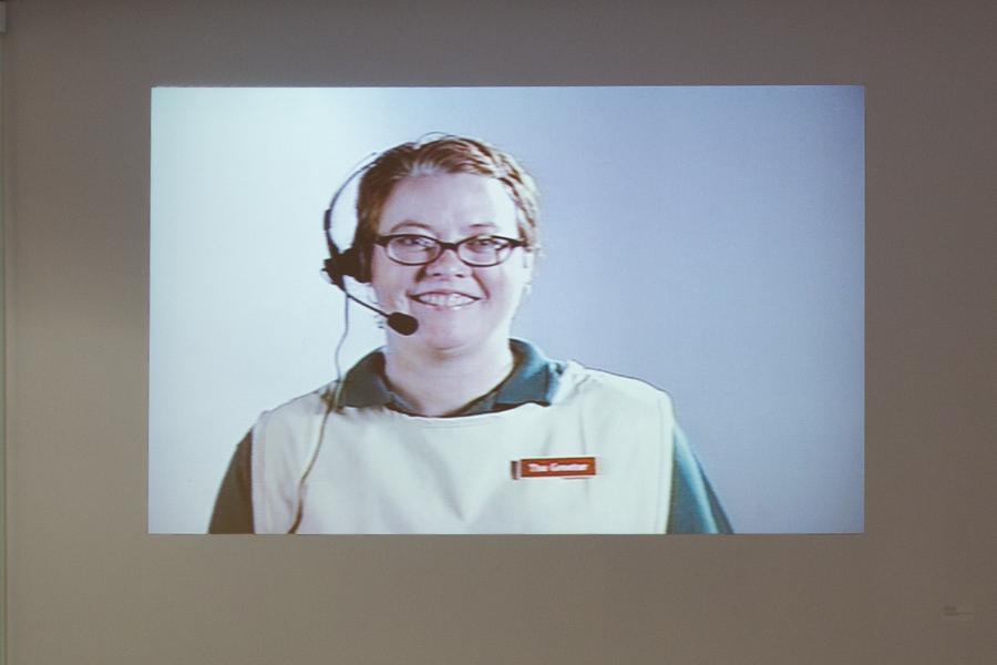 Projection of a woman with glasses and a headset smiling, labeled "The Greeter."