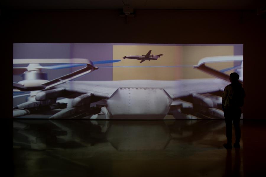 A gallery projection featuring close-up imagery of drones and aircraft, with a silhouetted viewer observing the installation.