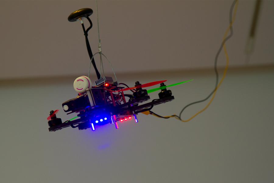 A suspended drone sculpture with colorful LED lights and visible wires in a gallery setting.