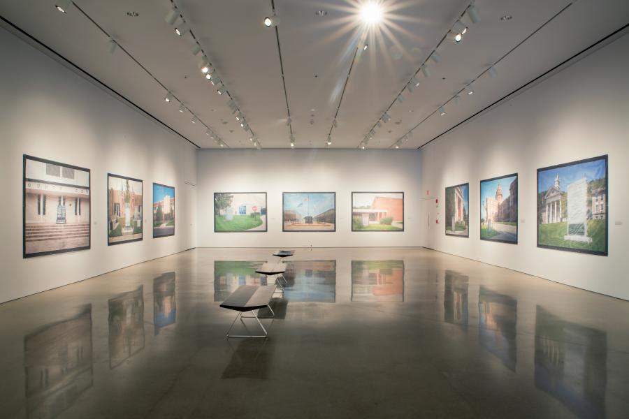 Gallery installation view featuring large-scale photographs of various courthouse buildings displayed on white walls, with polished concrete floors reflecting the artworks and benches arranged for viewers.