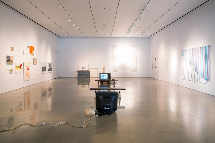 A contemporary gallery installation with mixed-media works, paintings, and a central display featuring an old television and benches.