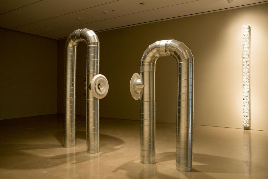A side view of the metallic pipe sculptures and a vertical wall installation in the dimly lit gallery.