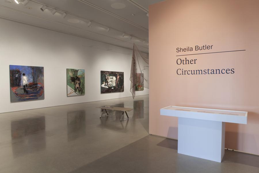 An installation view of Sheila Butler's exhibition "Other Circumstances" at the School of Art Gallery, showcasing a blend of large-scale narrative paintings and delicate hanging textiles, set against a muted peach title wall that introduces the exhibition.