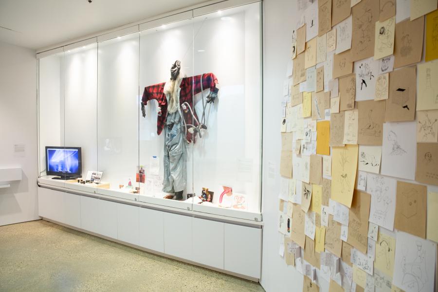 A glass display case featuring a mannequin dressed in mixed-media art and surrounded by sketches, objects, and photographs.