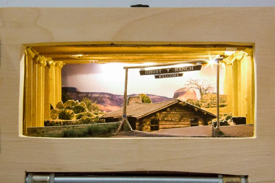 Close-up of the illuminated diorama inside "Georgia O’Keefe Country," showing a detailed miniature landscape with a wooden building and "Ghost Ranch Welcome" sign.