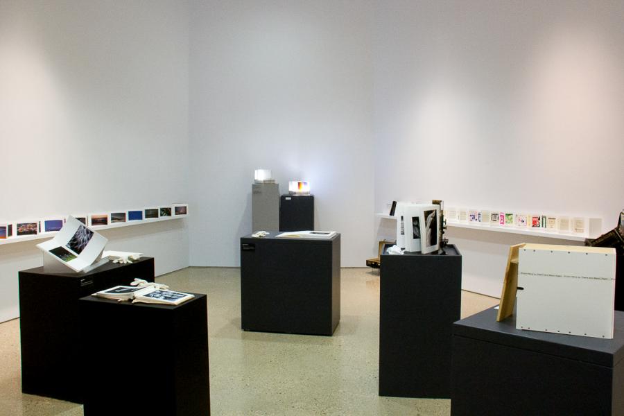 Gallery view with multiple black pedestals displaying Erwin Huebner’s artist books. Shelves along the wall hold additional works, including a line of colorful photographs.